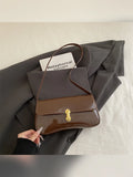 Bolso de hombro café brillante con cierre dorado sobre ropa gris