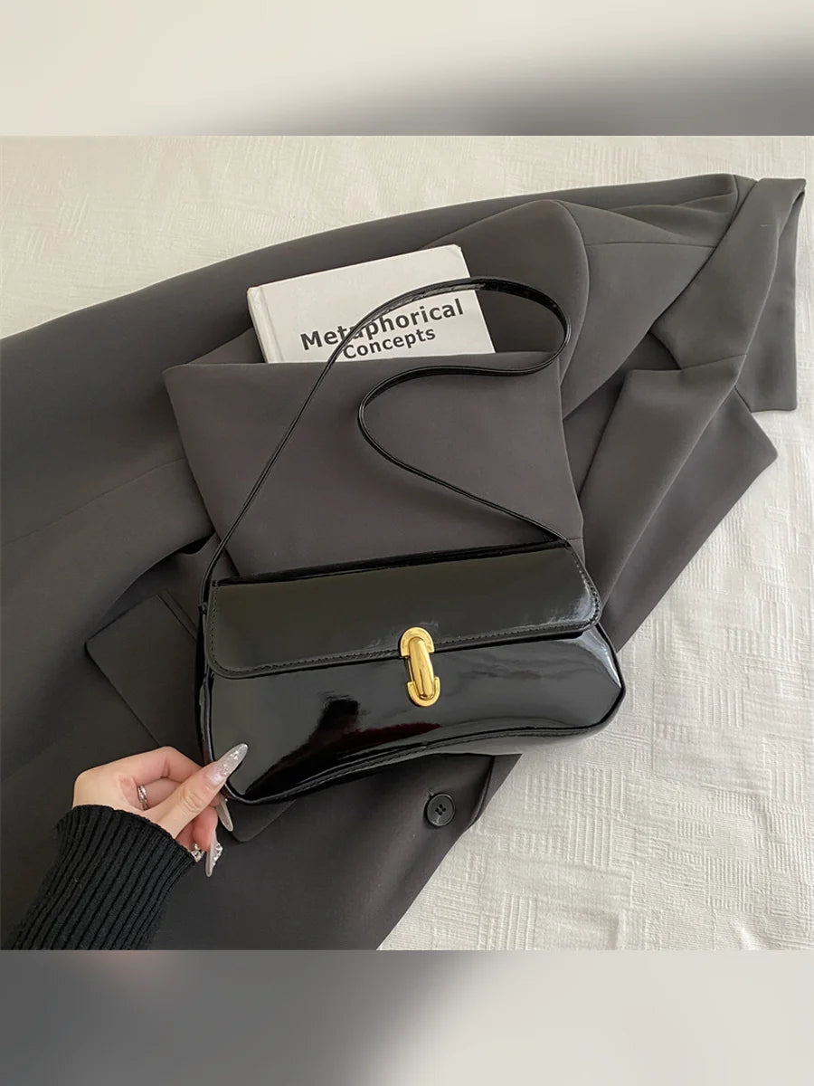 Bolso de hombro negro brillante con cierre dorado sobre ropa gris